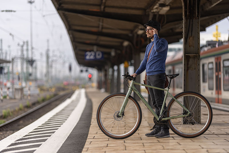 mann-mit-fahrrad-am-bahngleis Ein Hipster-Tpy-Mann steht mit seinem olivgrünen Stadtrad an einem Bahngleis und hält sich am Kopf. Er trägt einen Schnauzbart und einen Helm.