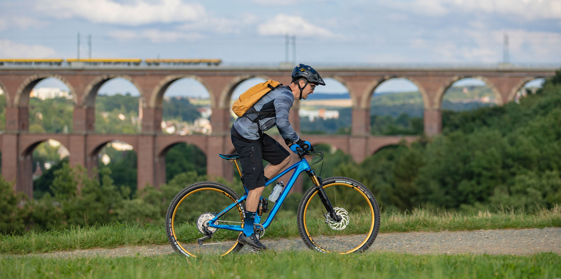 radfahrer-mountainbike-aquaedukt Ein Mountainbiker mit gelbem Rucksack fährt vor Landschaft stehend auf seinem Rad. Im Hintergrund ist groß ein Viadukt zu sehen.
