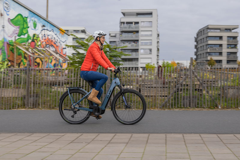 radfahrerin-im-urbanen-raum-auf-ebike Eine Radfahrerin im Rentner-Alter fährt auf einem grauen E-Bike der Marke Cube durch ein Stadtgebiet. im Hintergrund sind Hochhäuser, Gärten und eine mit bunten Graffiti besprühte Wand.